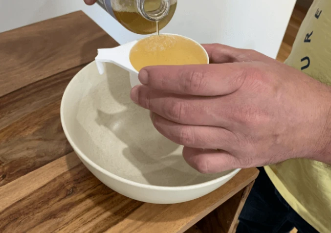 Pouring liquid castile soap from a bottle into a measuring cup over a mixing bowl.